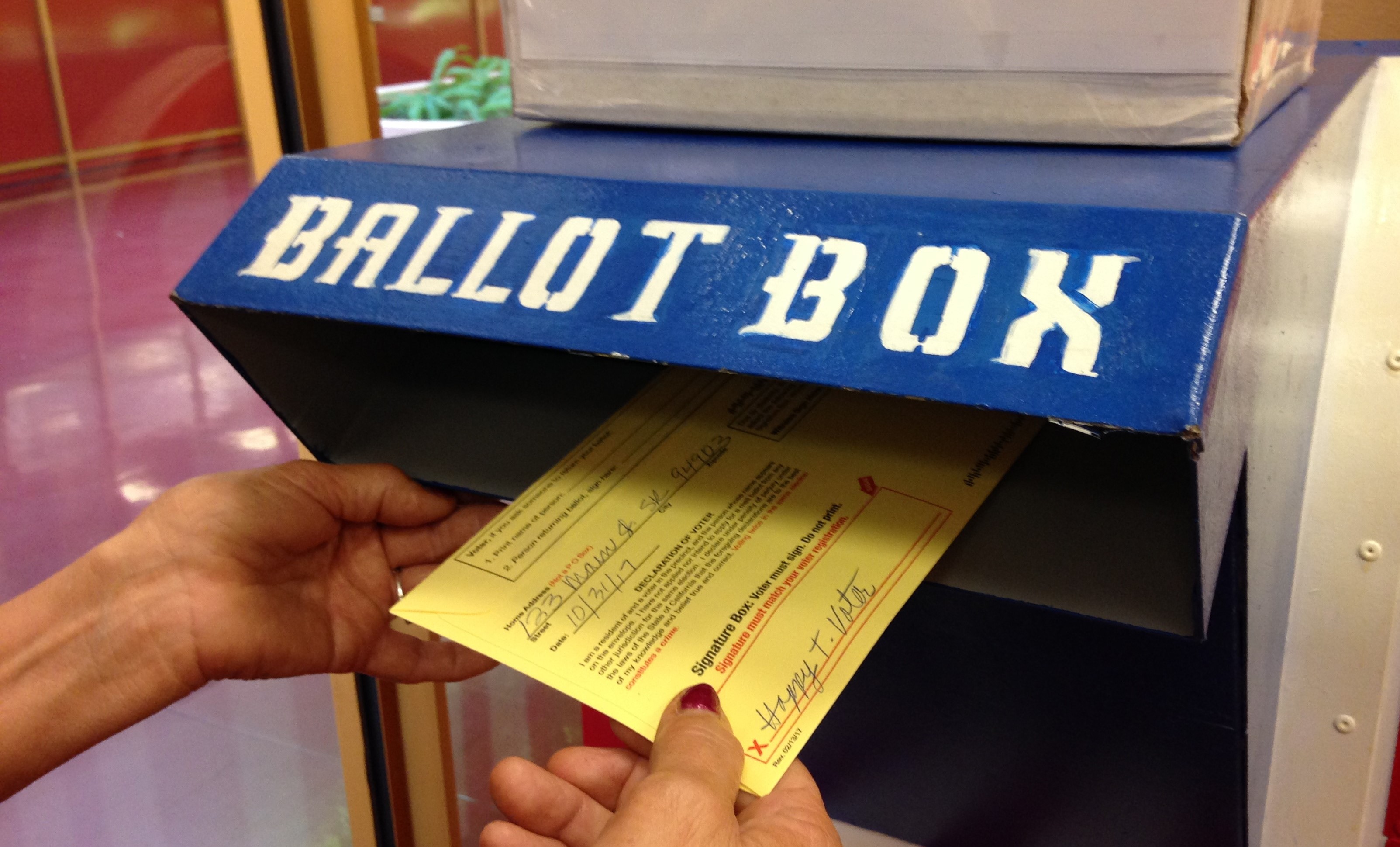 A closeup view of a person's hand dropping a ballot envelope in a Elections dropbox.