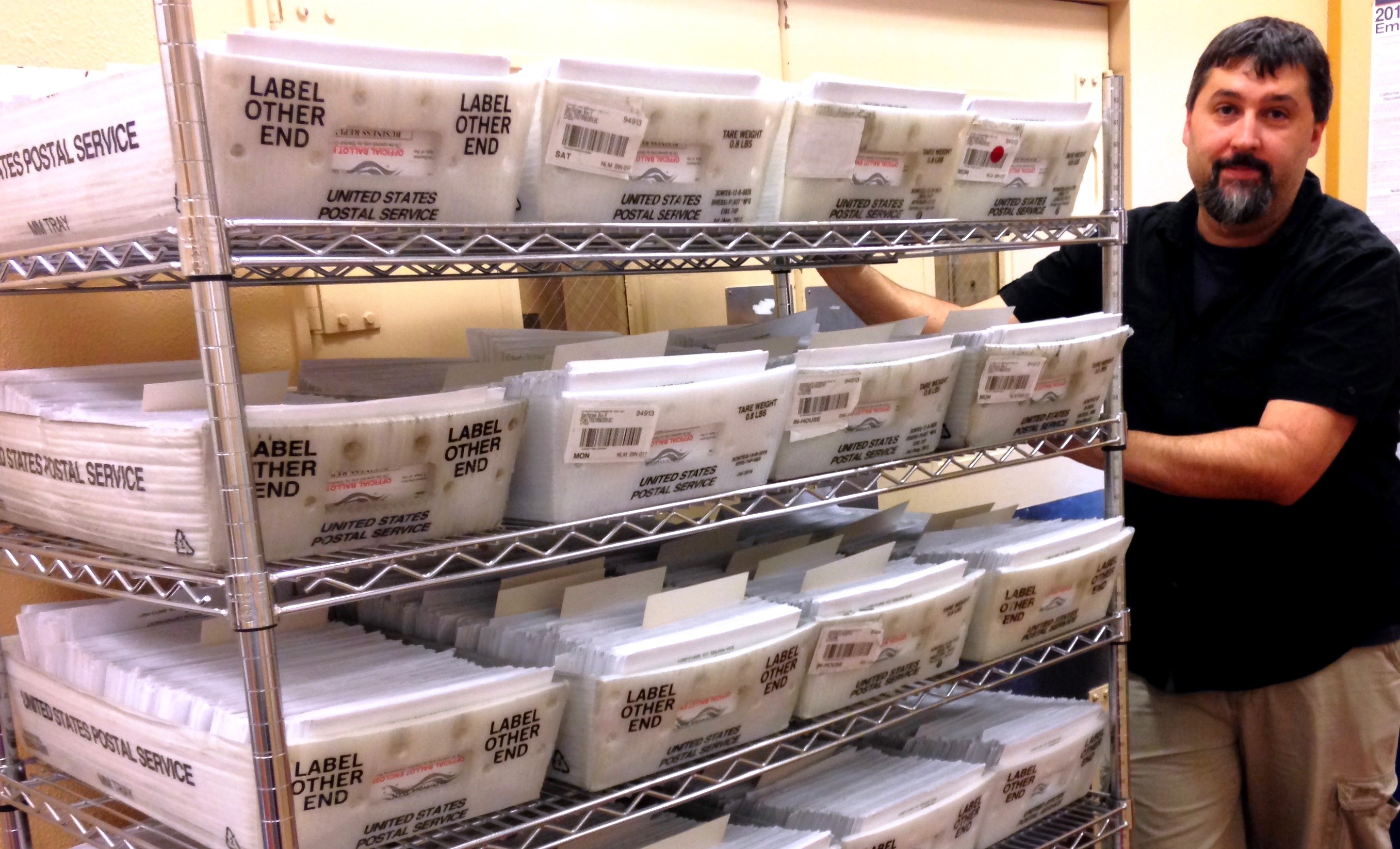 A male elections worker holds a mobile rack that holds thousands of ballot envelopes.