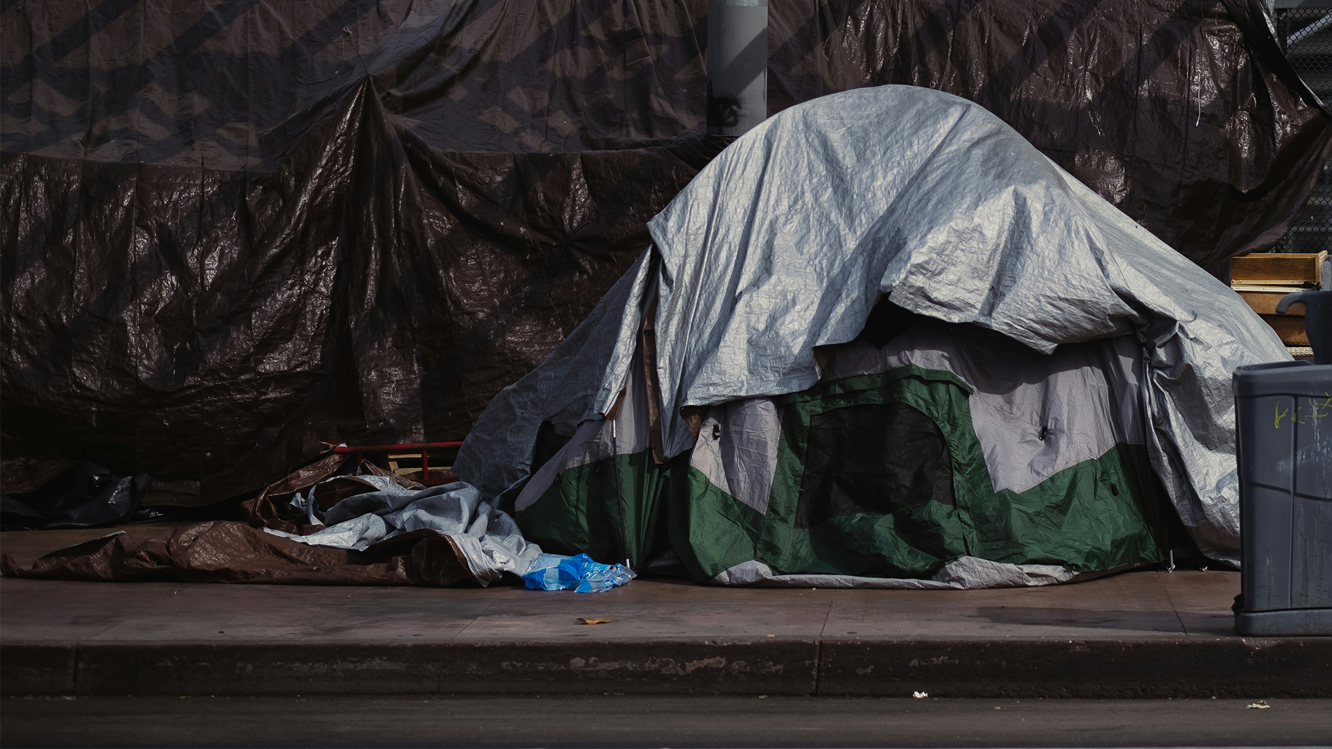 A homeless camp, with a green and silver tent and black tarp are set up along a sidewalk.