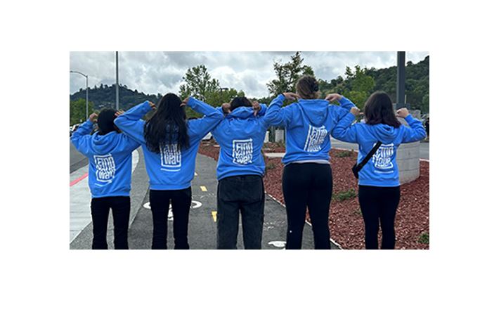 Five young people stand with their backs to the camera while wearing 9 to 25 sweatshirts.