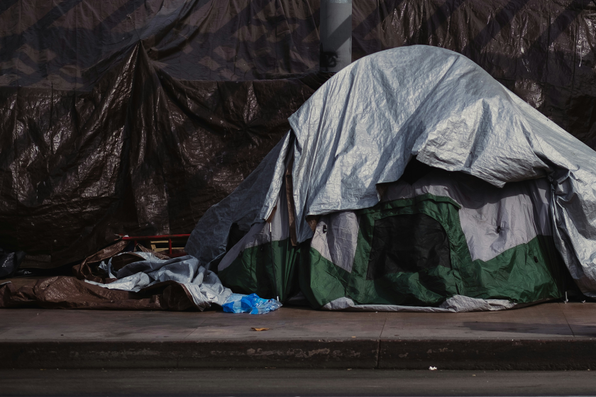A homeless camp, with a green and silver tent and black tarp are set up along a sidewalk.
